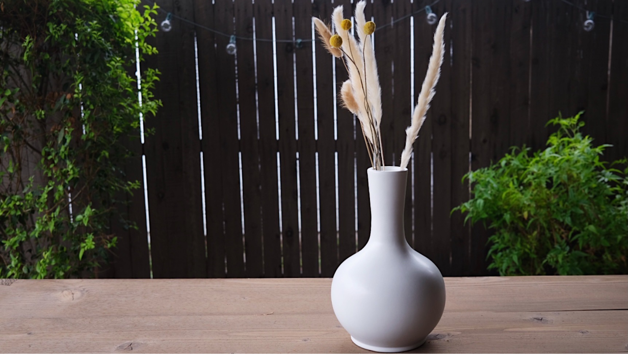 The white, medium sized urn sitting outside on a wooden dining table surrounded by green landscape